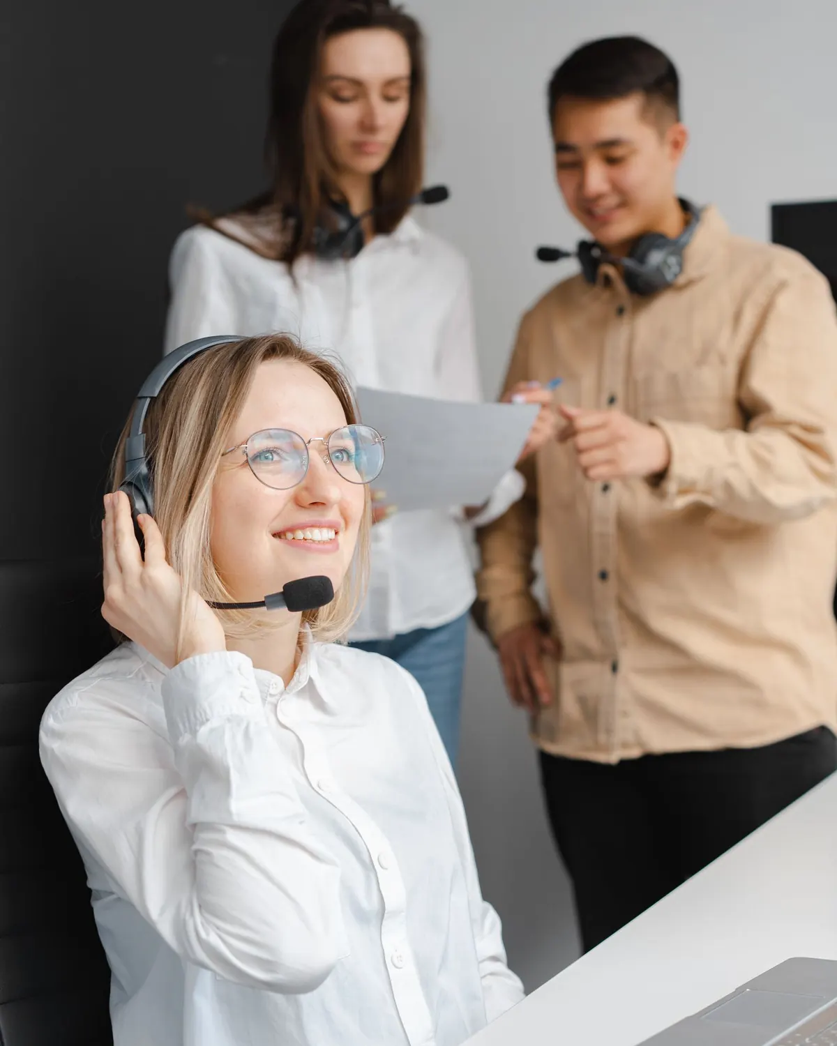Atendente sorrindo com headset na mesa de trabalho, com dois colegas ao fundo